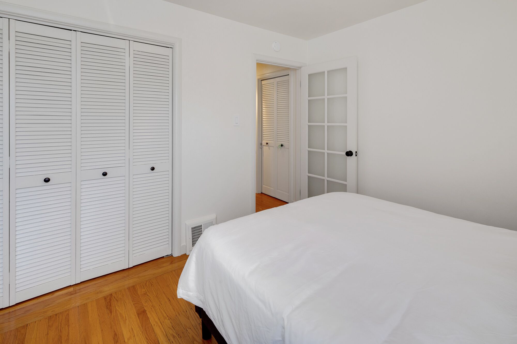 Un dormitorio con cama, piso de madera, paredes blancas y dos puertas con persianas a persiana.