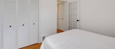Un dormitorio con cama, piso de madera, paredes blancas y dos puertas con persianas a persiana.