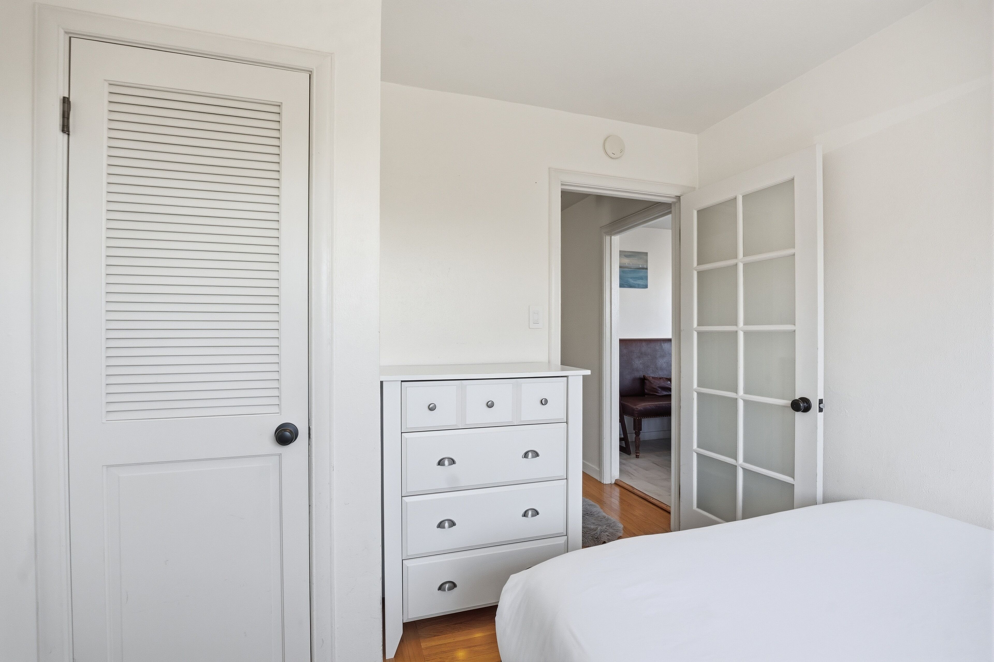 A bedroom with a white door, a dresser, a bed, and a glass door leading to another room.