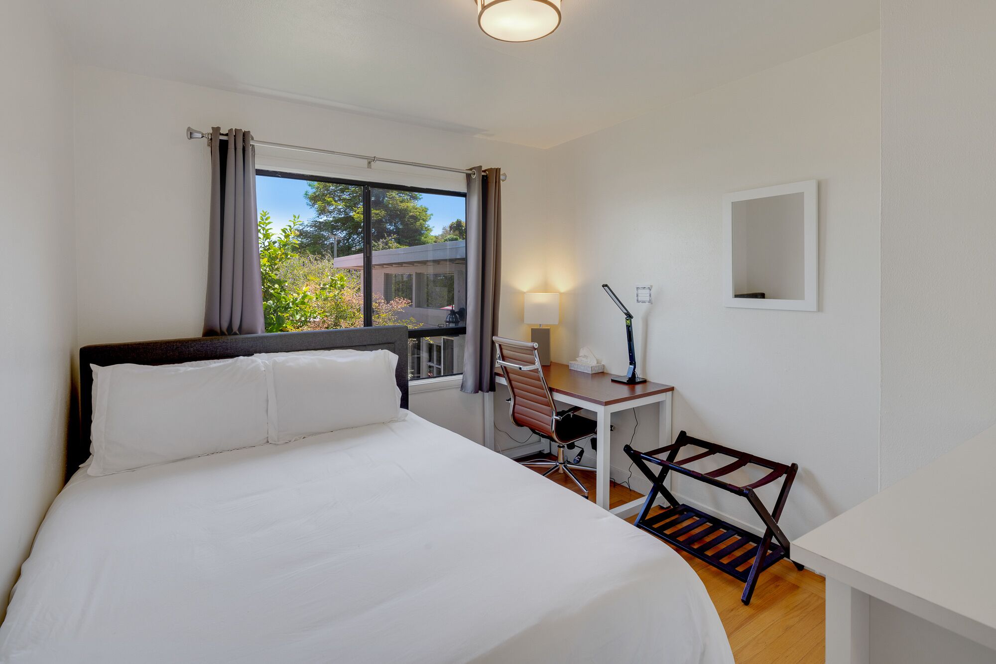 A hotel room with a bed, desk, chair, lamp, and a view of greenery outside.