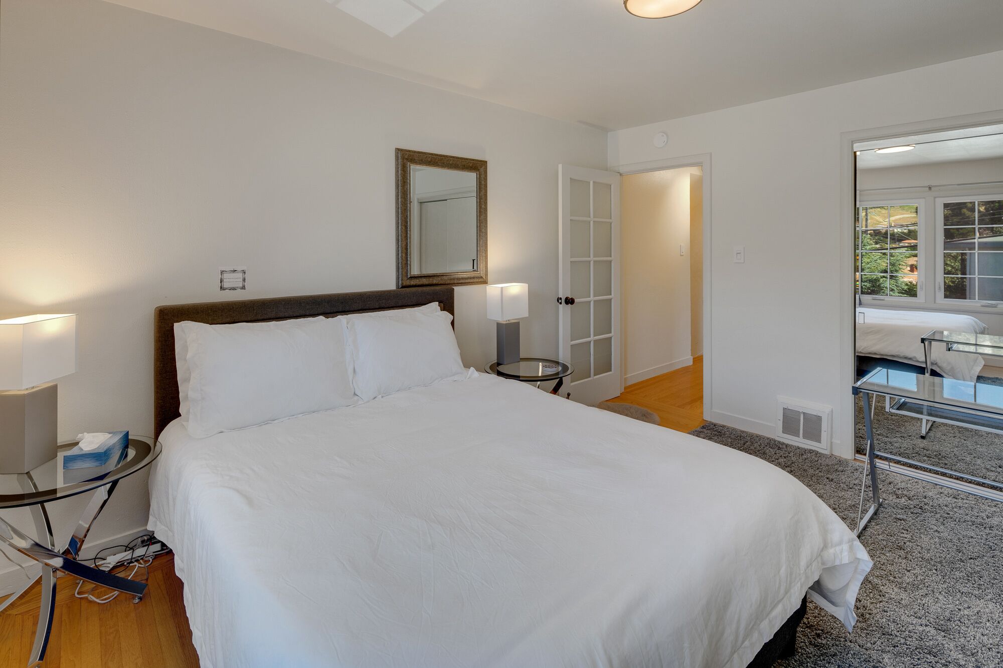 Een hotelkamer met een groot bed, bedlampjes, een spiegel, een deur naar een balkon en een kleine tafel met een lamp.