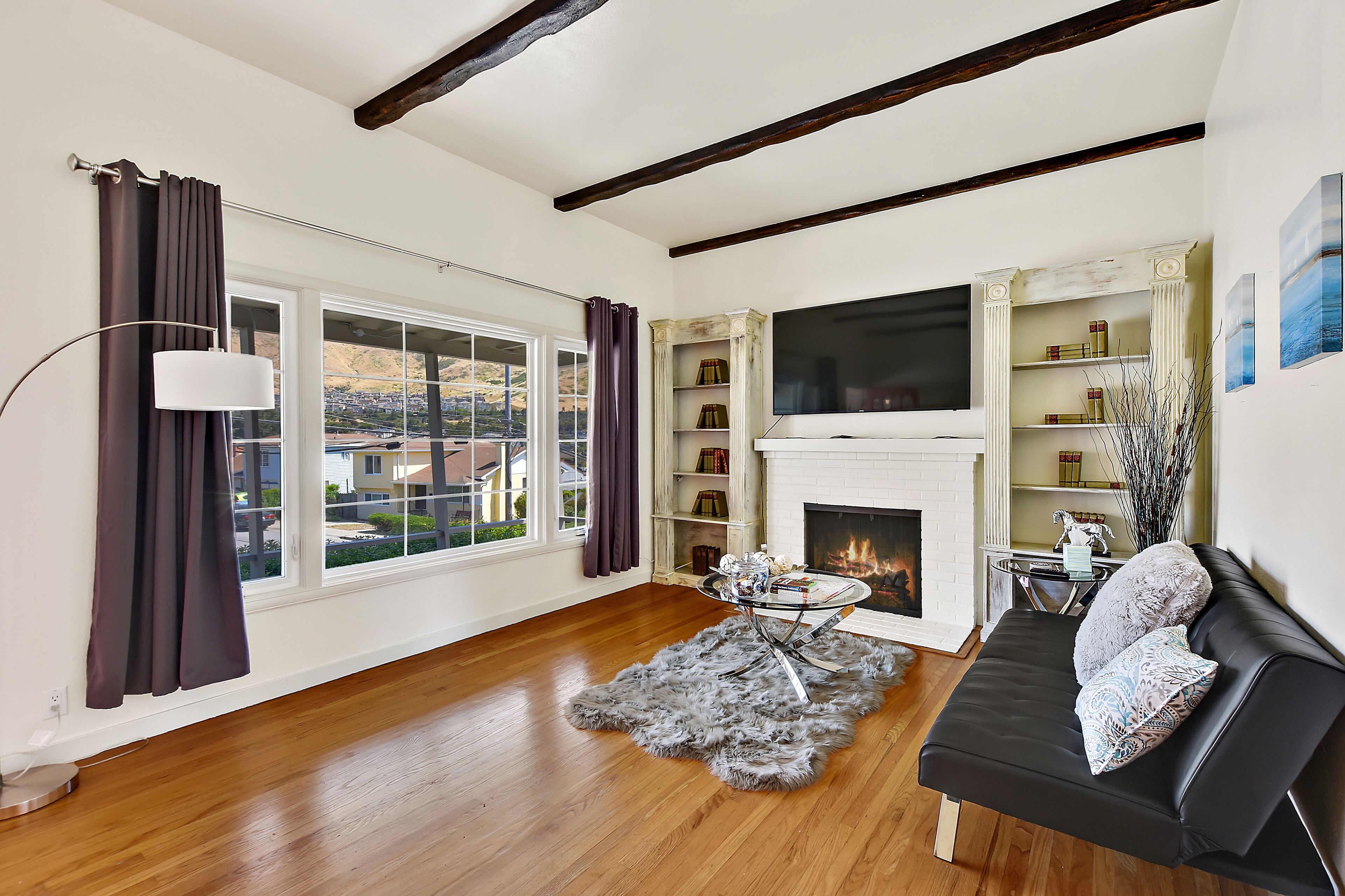 A living room with a fireplace, a black sofa, a glass coffee table, and a view of the outside.