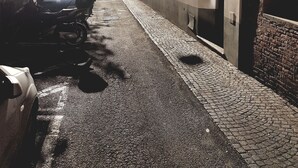 Calle empedrada estrecha flanqueada por edificios históricos, con una fila de coches aparcados y una puerta visible a la derecha.