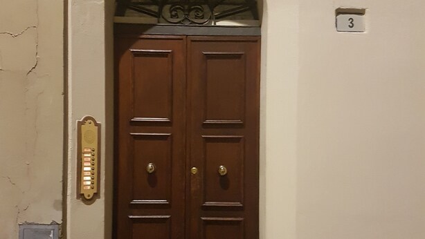 Una puerta doble de madera con un escalón que lleva a ella, un buzón a la izquierda y una placa con el número '3' en la pared a la derecha.