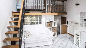 Un dormitorio tipo loft con escalera de madera, una cama litera, un sofá blanco y una pequeña cocina.