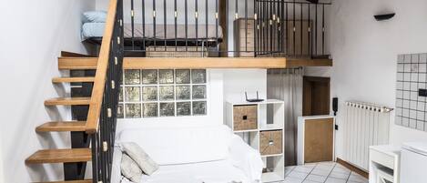 Quarto em estilo loft com escada de madeira, cama beliche, sofá branco e pequena cozinha.