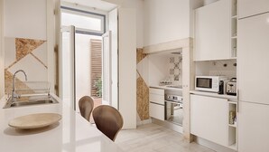 A modern kitchen with white cabinetry, a built-in oven, microwave, and a sink. There is a dining area with a wooden table and chairs. - Romantic Gem in Alfama (Lisboa)