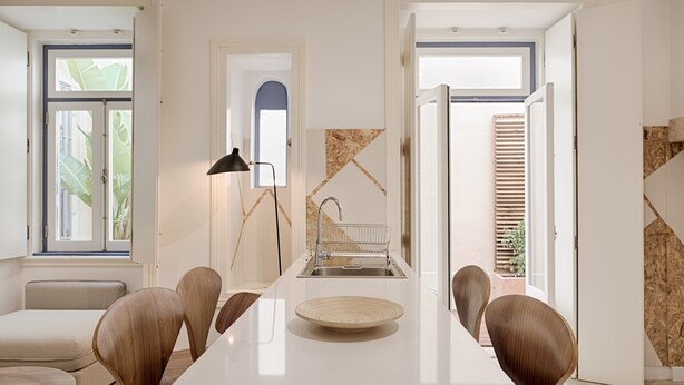 A modern kitchen with a white countertop, a round island, and a built-in sink. There are wooden chairs around the table, and a window with blinds is visible. - Romantic Gem in Alfama (Lisboa)