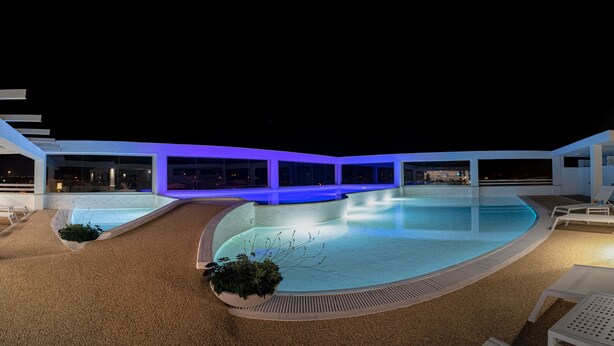A nighttime pool area with a curved design, surrounded by white lounge chairs and a covered walkway. - Stele di Nora (Pula)