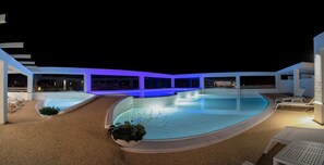 A nighttime pool area with a curved design, surrounded by white lounge chairs and a covered walkway.