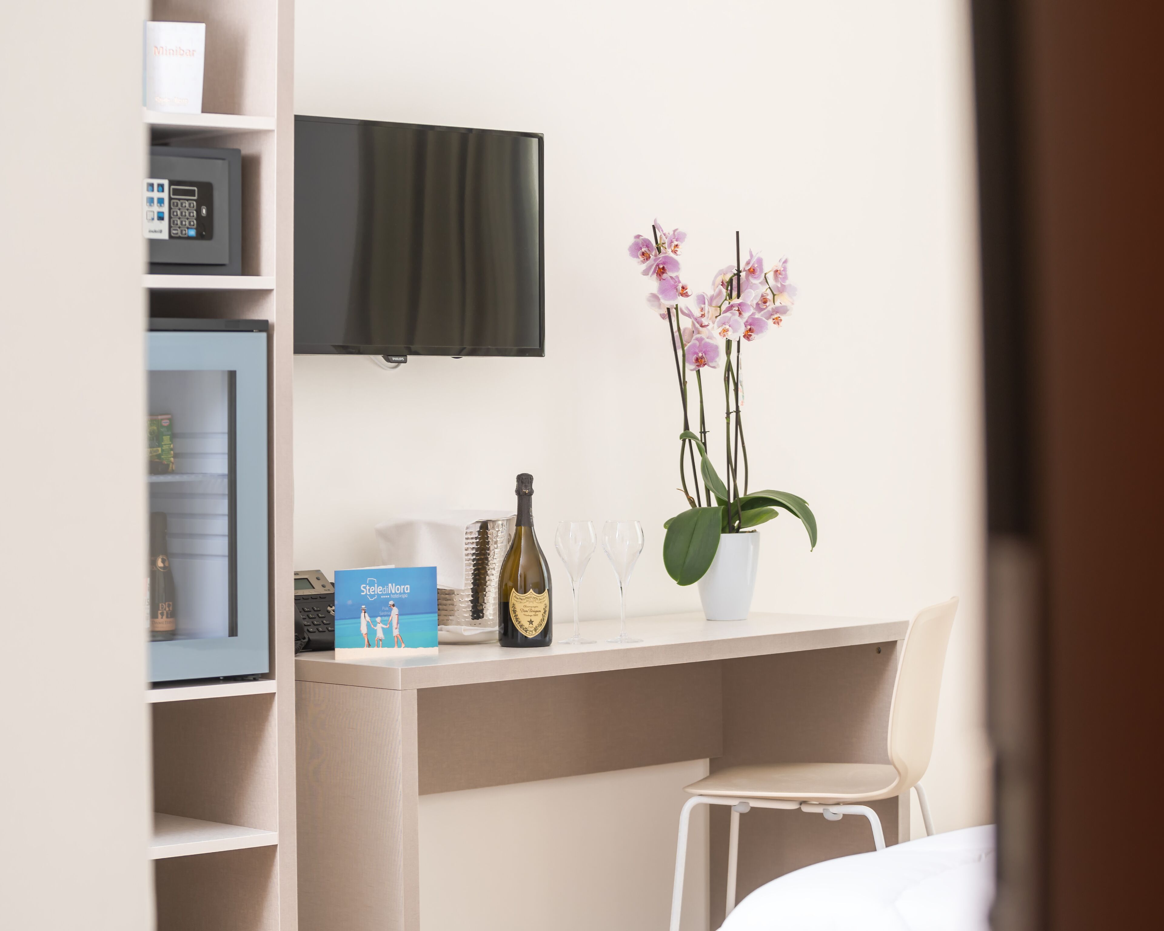 A hotel room with a desk, a television mounted on the wall, a potted orchid, a bottle of wine, and a couple of glasses.