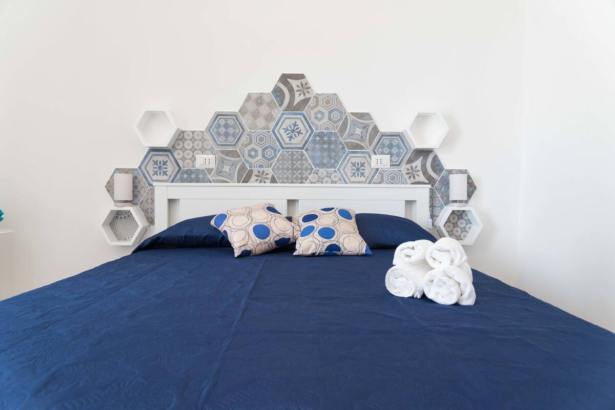 Una camera da letto con una coperta blu, due cuscini decorati, una testiera bianca e un muro rivestito di piastrelle esagonali.