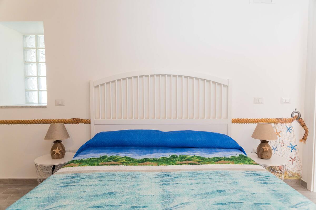 Camera da letto con un letto e una copriletto a tema oceano blu, una testiera bianca e due comodini con lampade.