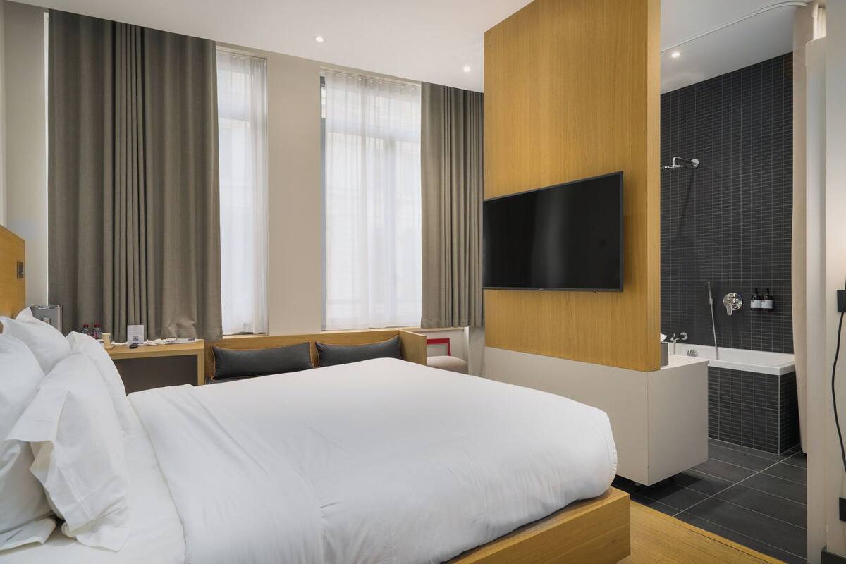 A modern hotel room with a large bed, a mounted TV, a bathroom with a bathtub, and a desk with a chair.