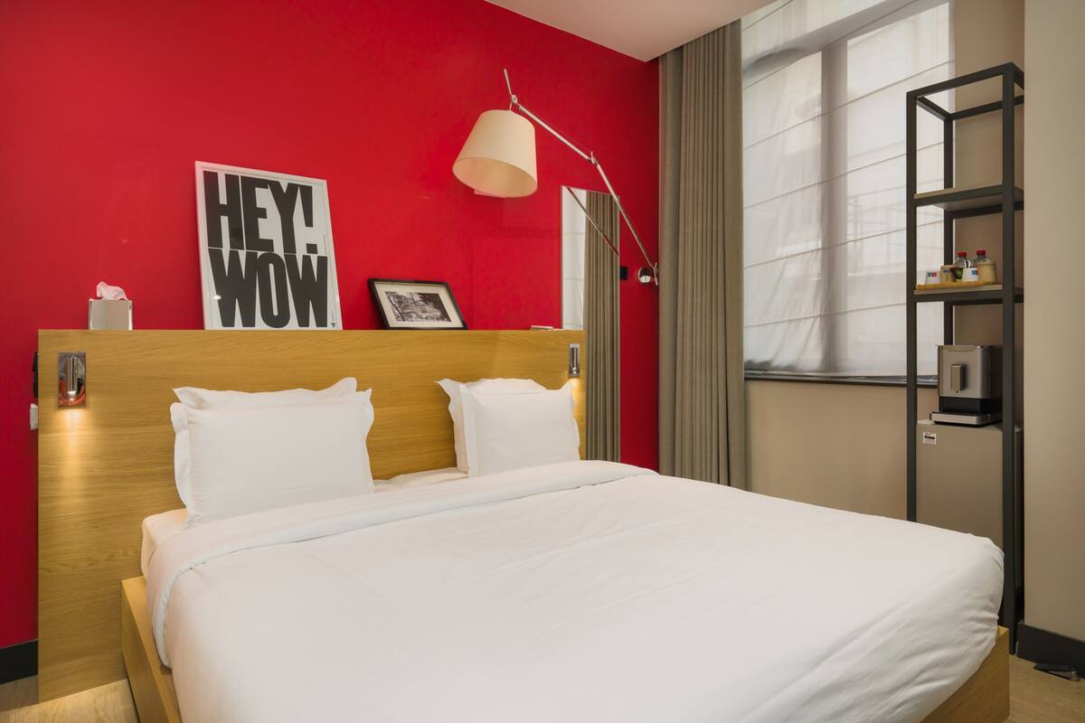 A hotel room with a large bed, a red accent wall, a framed picture, a bedside lamp, and a shelf with toiletries.