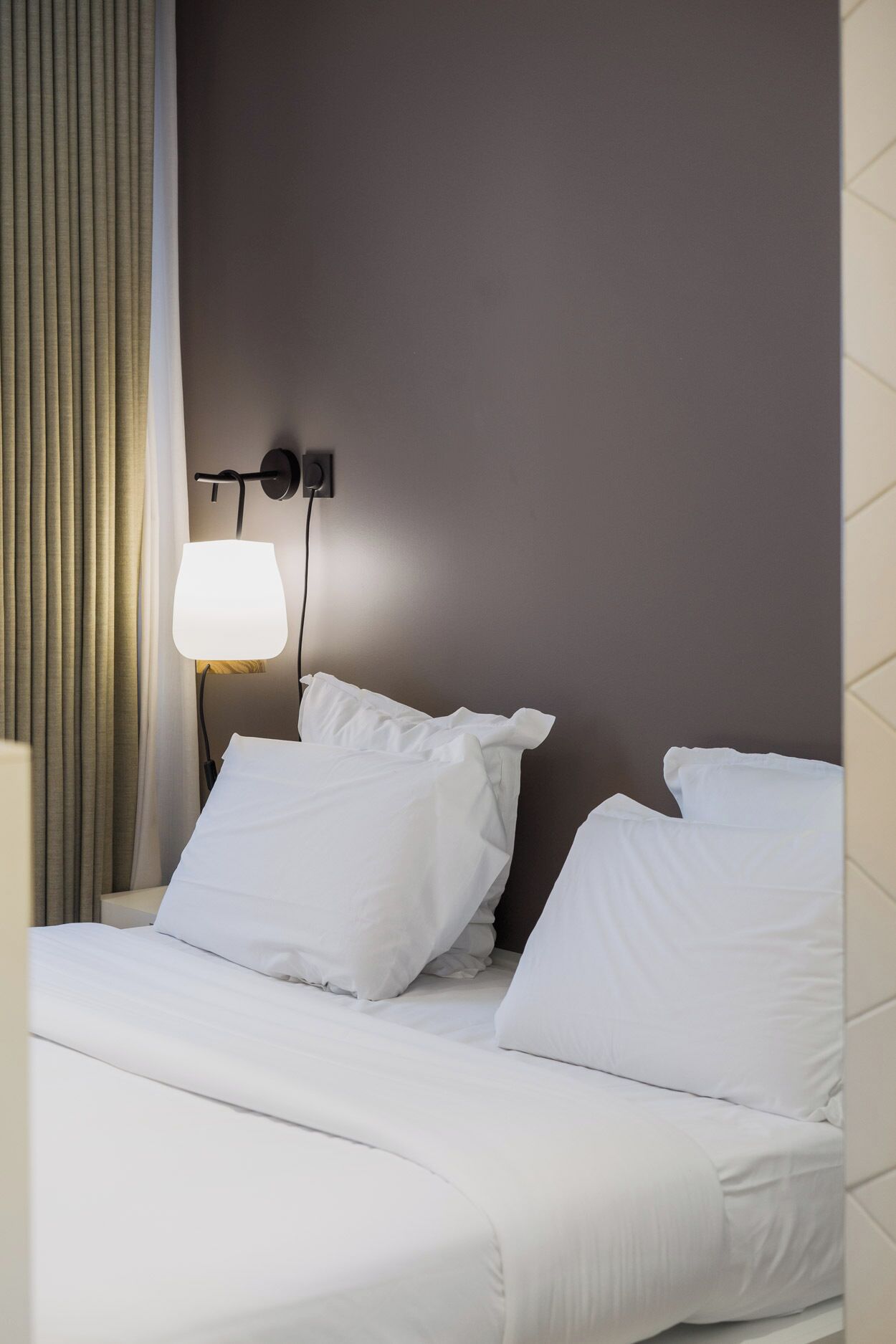 Ein Hotelzimmer mit Bett, weißen Laken, einer Wandlampe und einem Fenster mit Vorhängen.