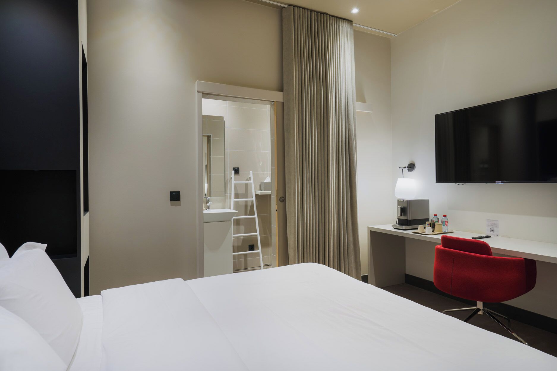 Habitación de hotel con una cama, un escritorio con ordenador, una silla roja, televisión y un baño visible a través de una puerta abierta.