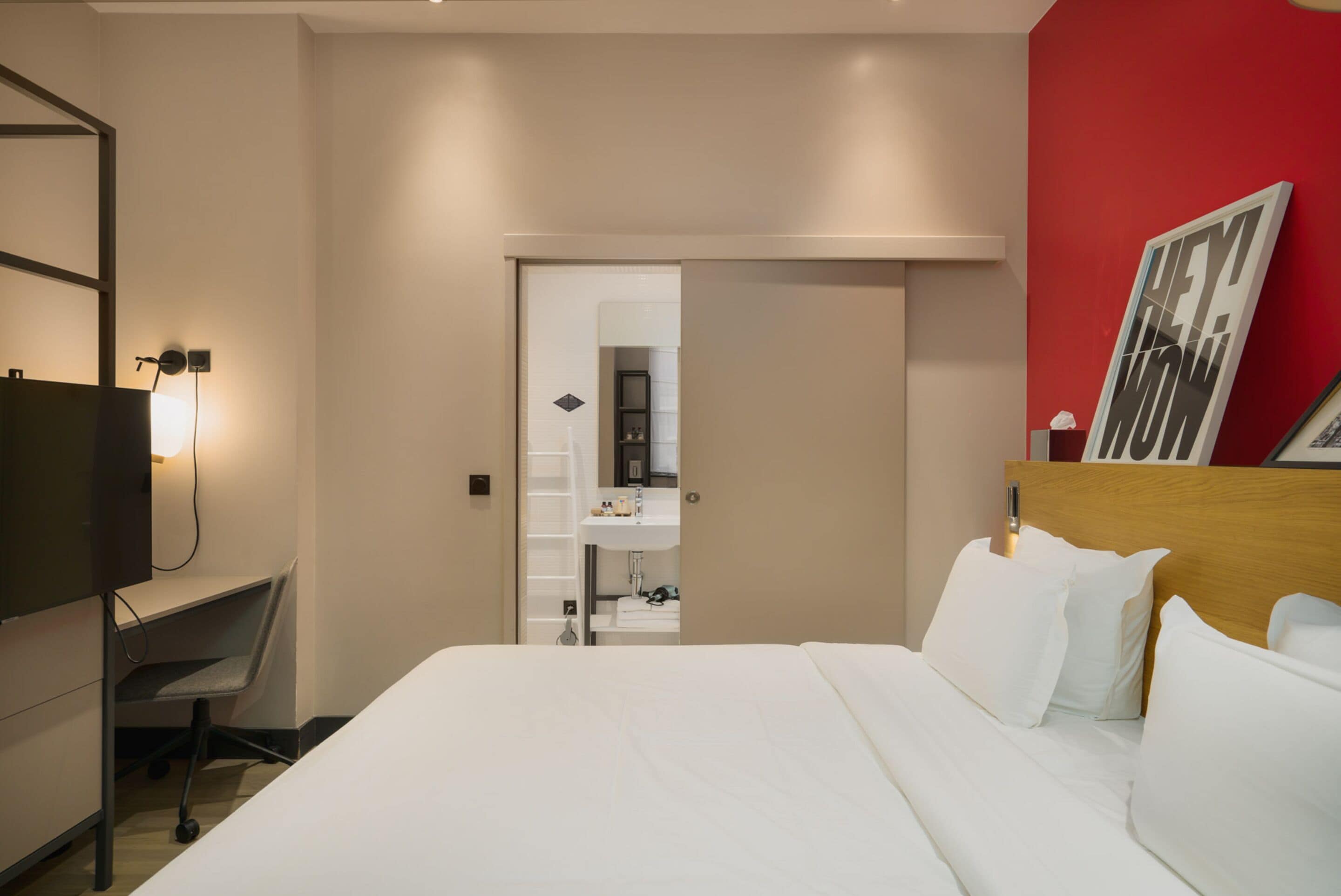 A hotel room with a bed, a desk, a TV, and a red accent wall.