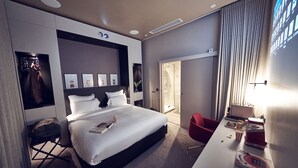 A modern hotel room with a large bed, a desk with a computer, a red chair, and a shower area. - Maison Nô (Lyon)
