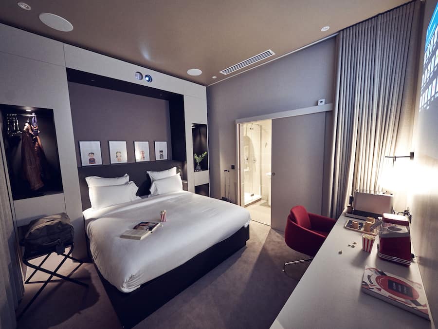 A modern hotel room with a large bed, a desk with a computer, a red chair, and a shower area.