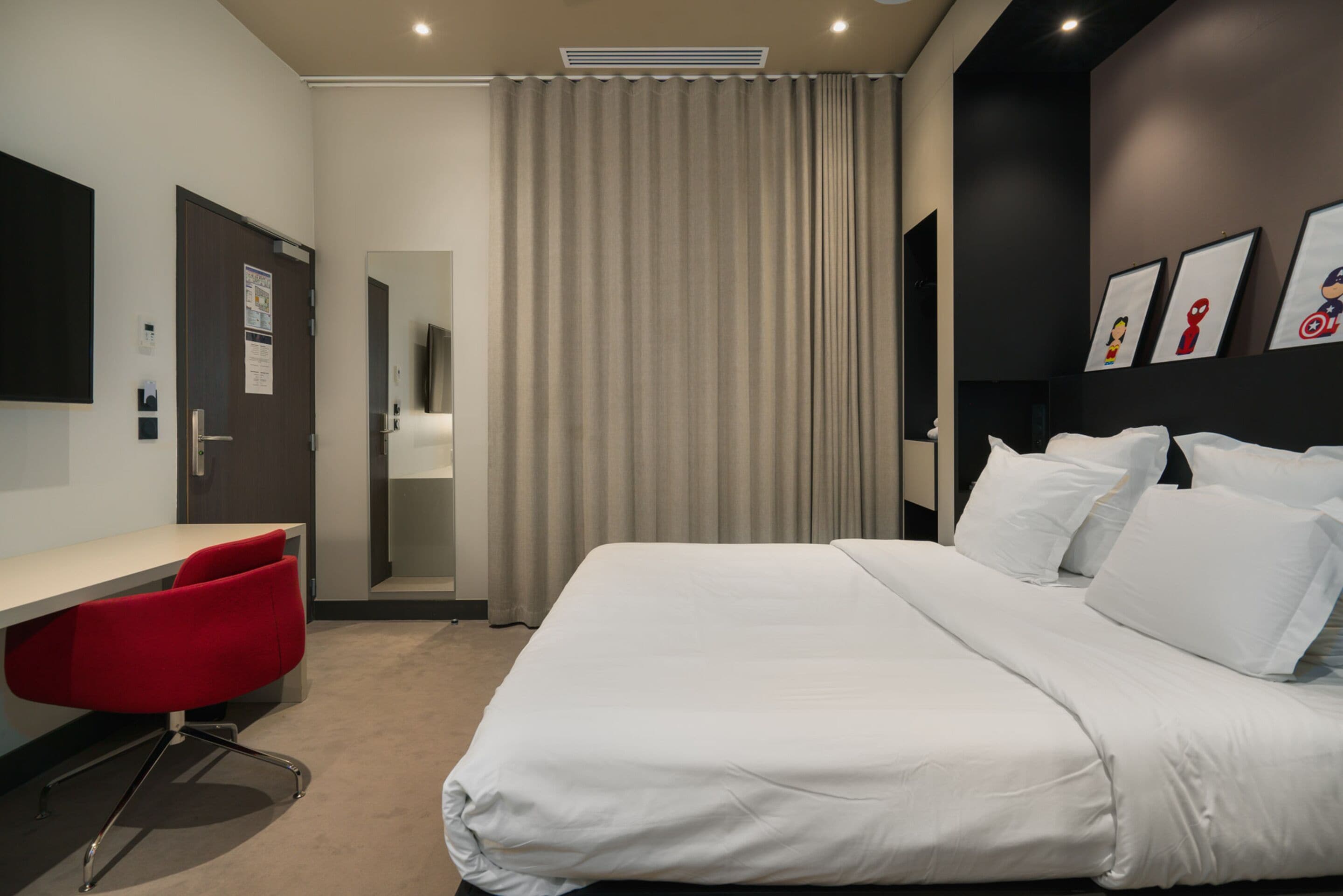 A modern hotel room with a large bed, a desk with a red chair, and a view of the hallway.