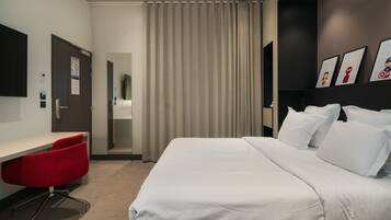 A modern hotel room with a large bed, a desk with a red chair, and a view of the hallway.