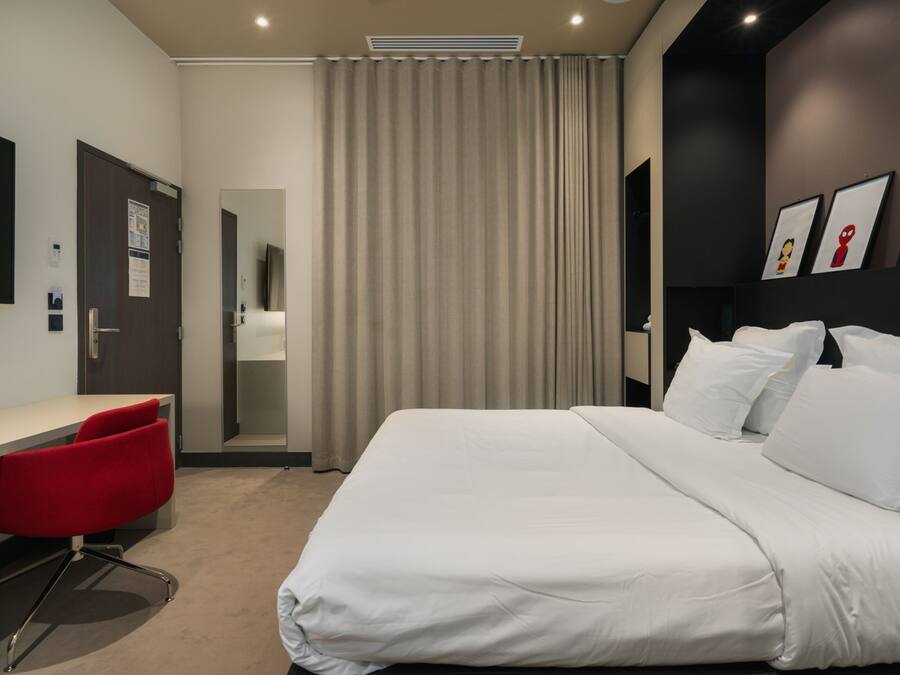 A modern hotel room with a large bed, a desk with a red chair, and a view of the hallway.