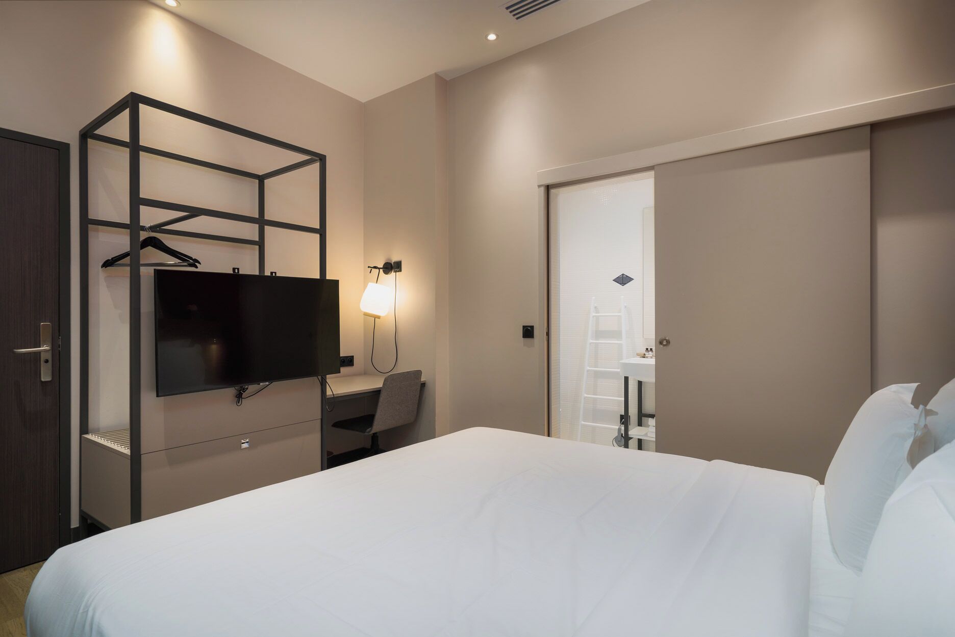 A hotel room with a bed, a desk with a chair, a TV mounted on a wall, a wardrobe, and a bathroom visible through an open door.
