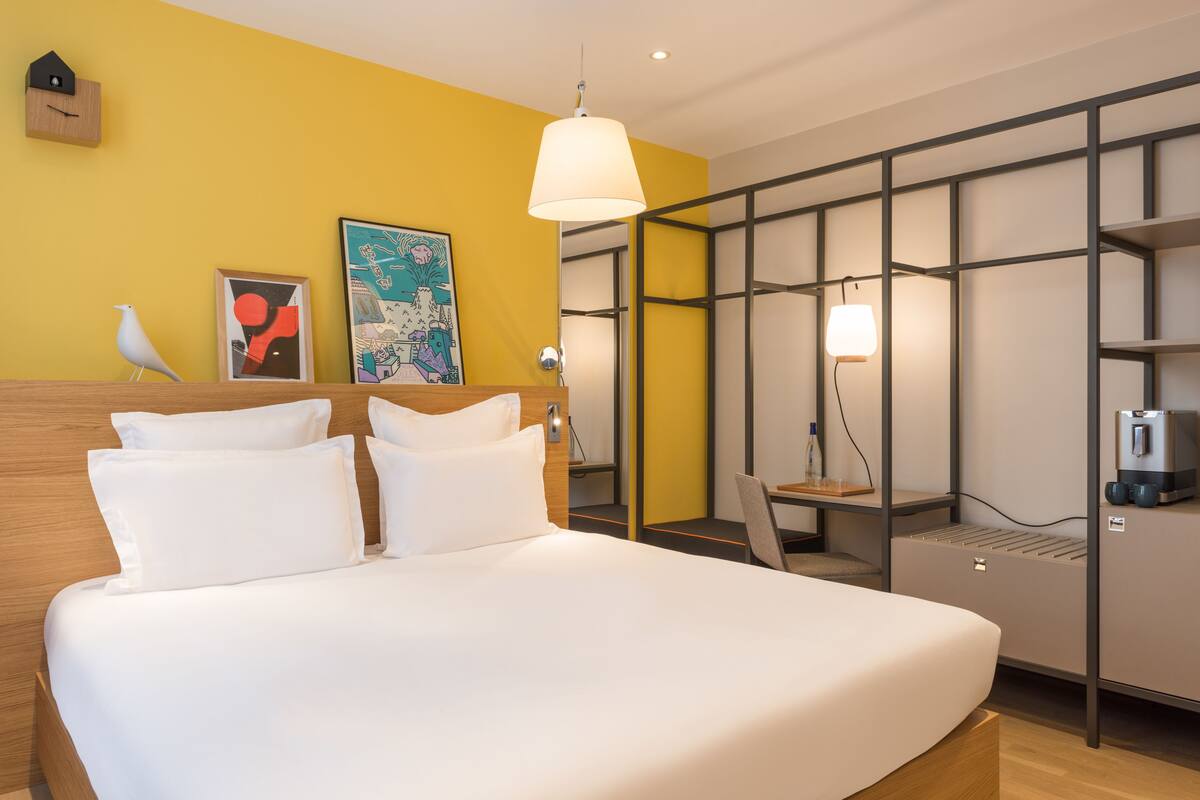 A hotel room with a bed, a desk, a chair, a lamp, and artwork on the wall.