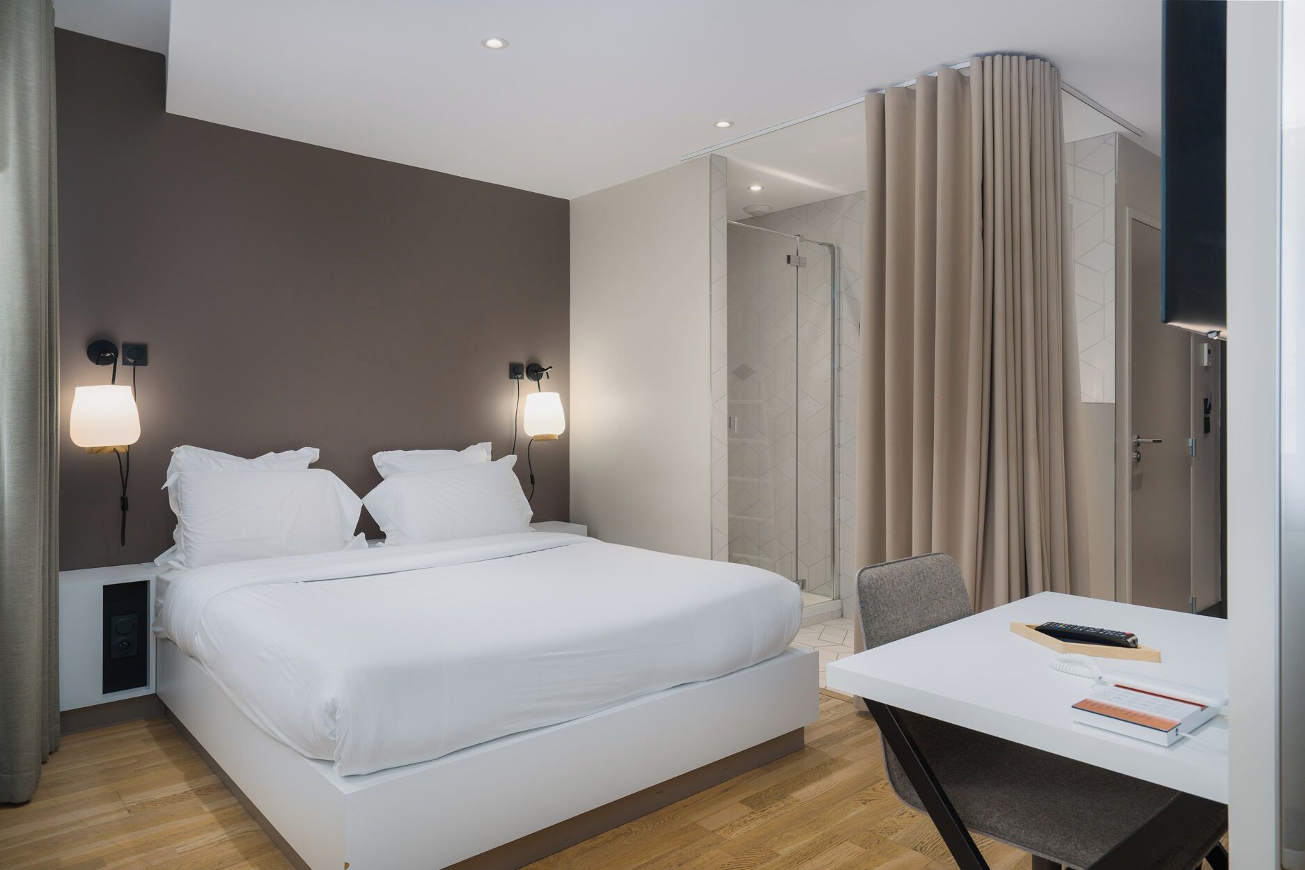 A modern hotel room with a large bed, bedside lamps, a shower, a desk with a chair, and a TV.