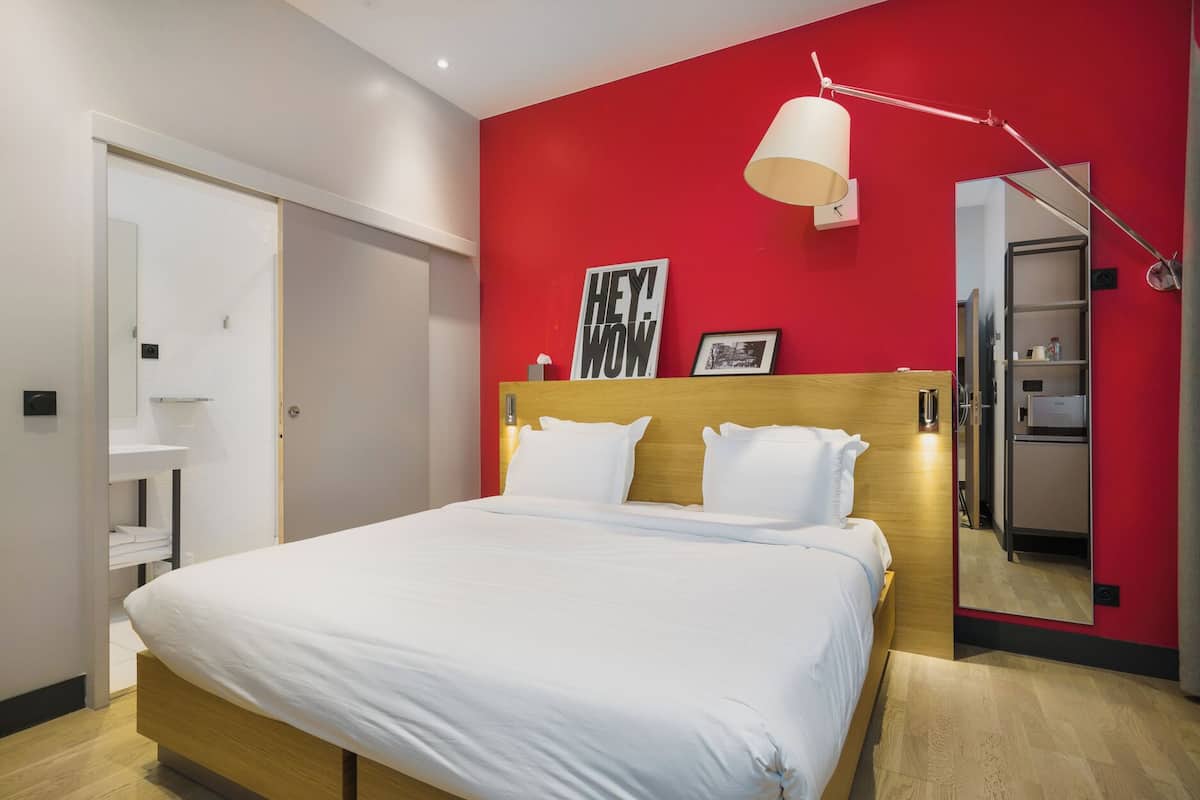 A hotel room with a bed, a red accent wall, a bedside lamp, a mirror, and a door.