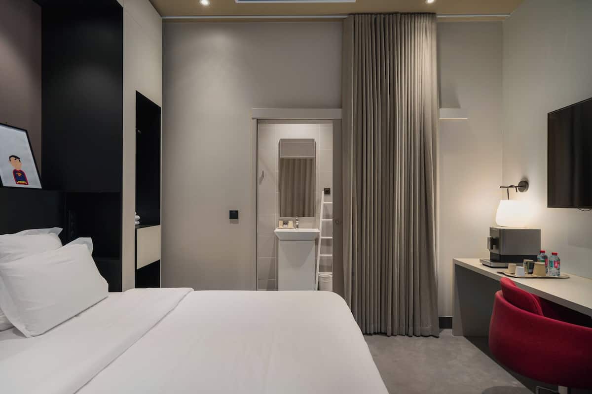 A hotel room with a bed, a desk with a lamp, a mirror, and a bathroom visible through a door.