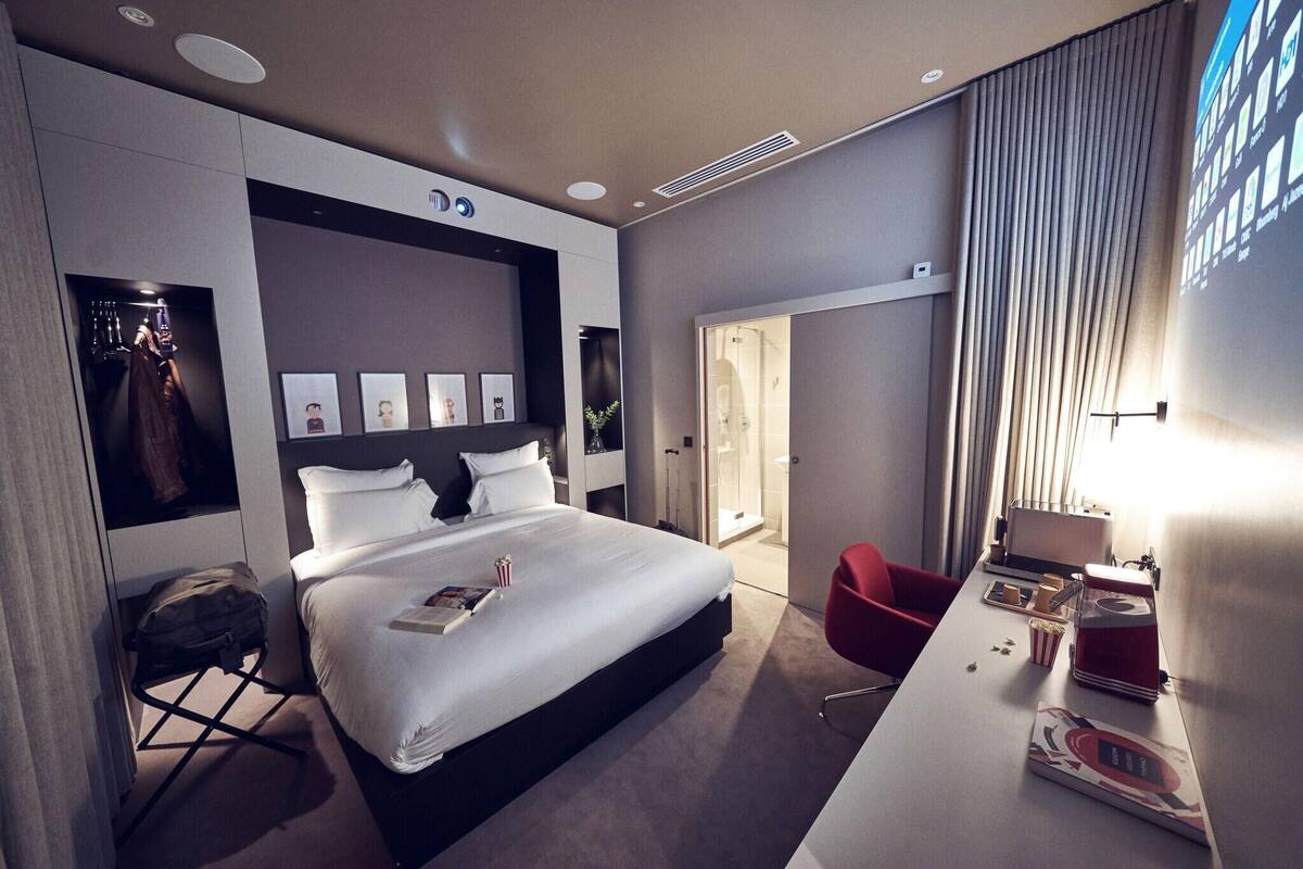 A modern hotel room with a large bed, a desk with a computer, a red chair, and a shower area.