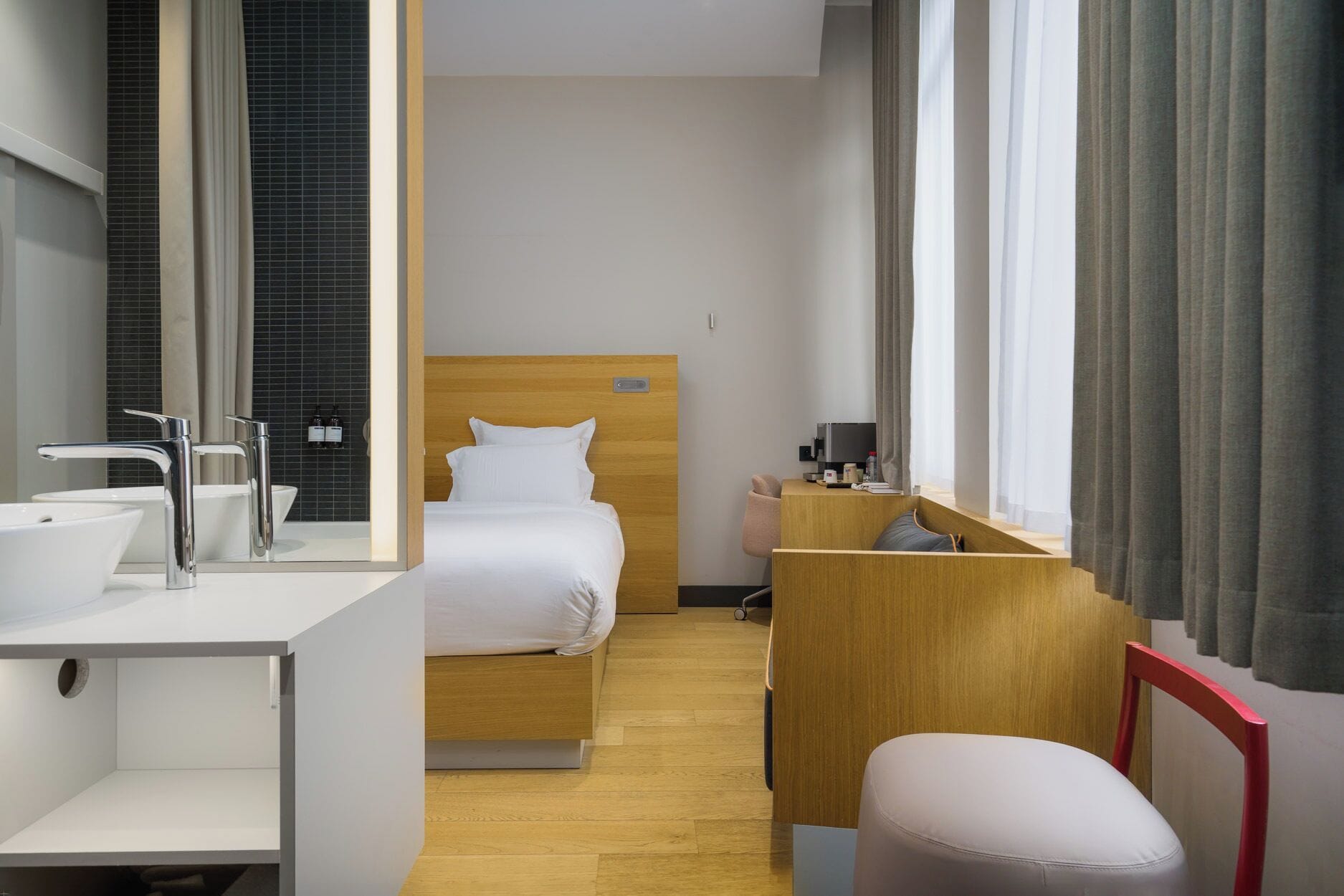 A hotel room with a wooden bed, a desk with a chair, a bathroom with a sink and mirror, and a large window with curtains.