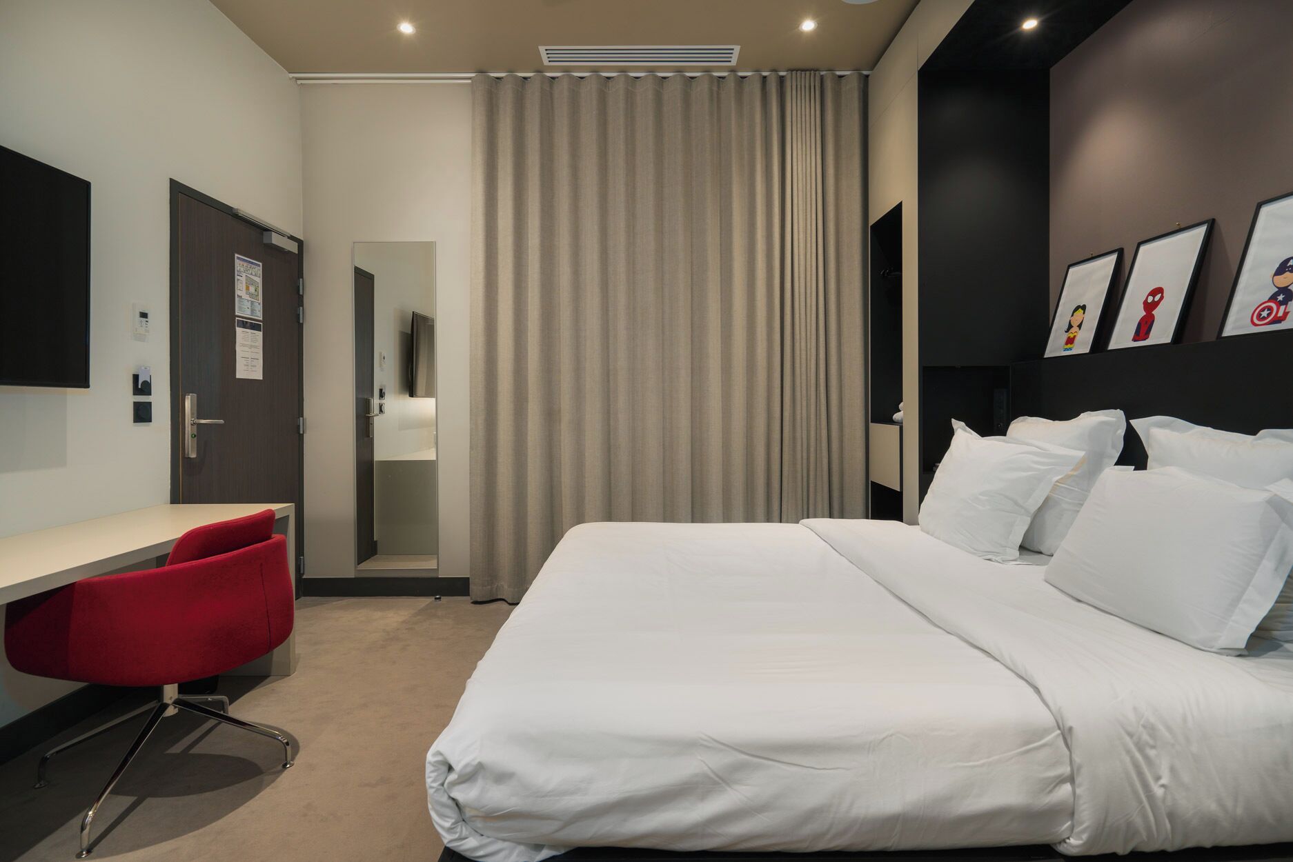 Una habitación de hotel con una cama, un escritorio, una silla roja, un espejo y una cortina.