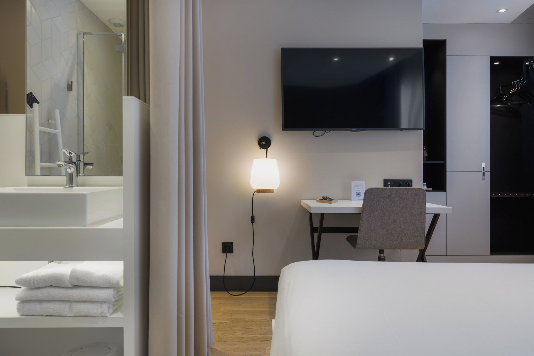 A modern hotel room with a large bed, a desk with a chair, a flat-screen TV mounted on the wall, a bathroom with a sink and mirror, and a bedside table with towels.