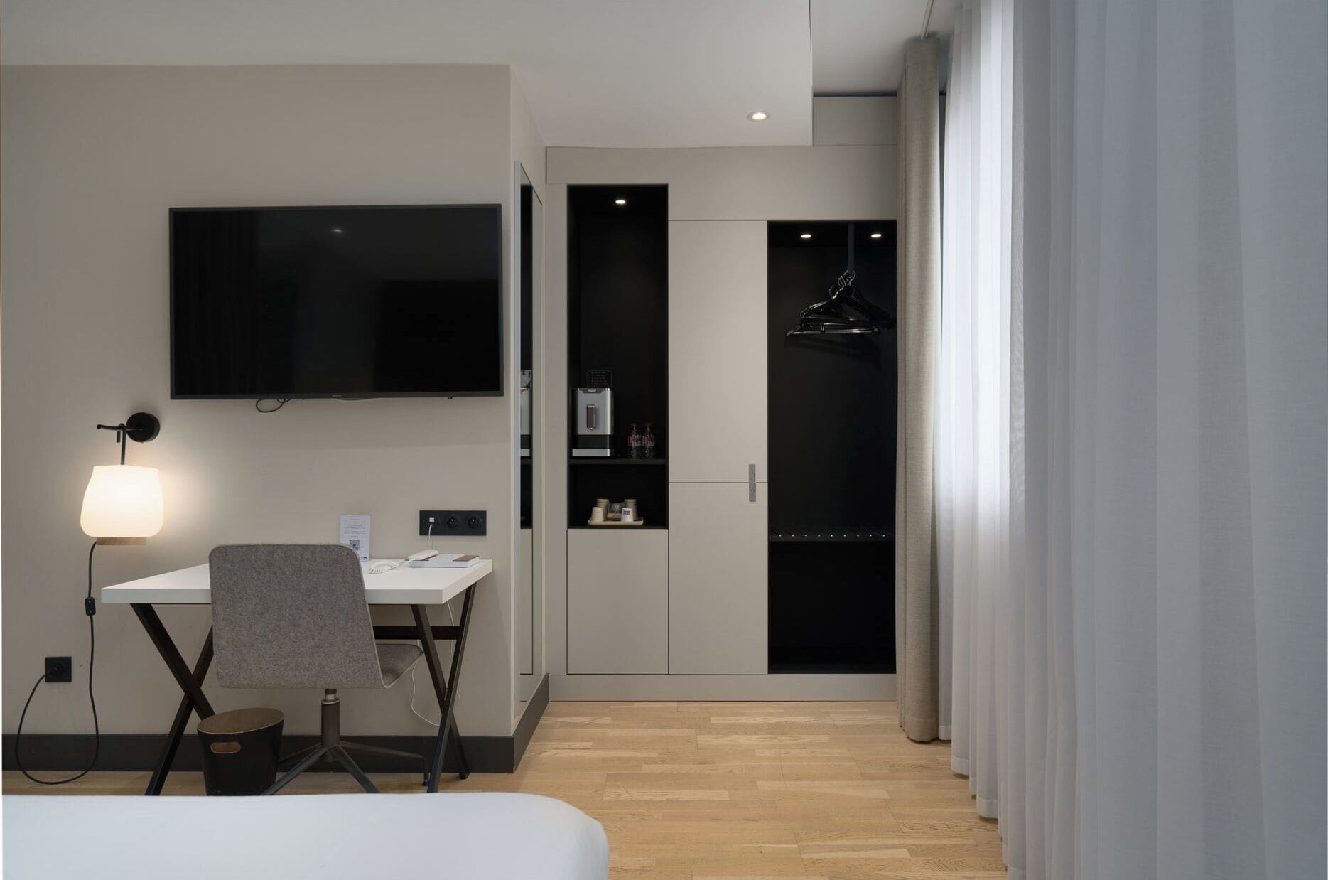 A modern hotel room with a desk, chair, flat-screen TV, wardrobe, and a bed.