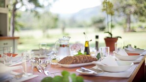 Una mesa de comedor dispuesta con platos, vasos y una hogaza de pan, con vistas a la vegetación y montañas al fondo.