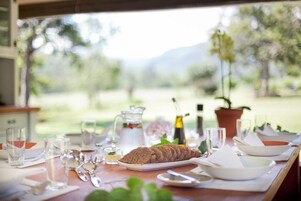 Una mesa de comedor dispuesta con platos, vasos y una hogaza de pan, con vistas a la vegetación y montañas al fondo.