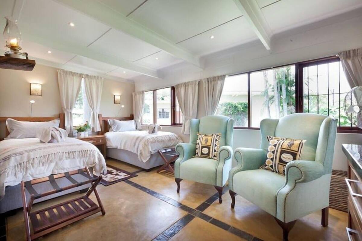 Een slaapkamer met twee bedden, een houten bankje en twee lichtblauwe fauteuils met gestreepte kussens.