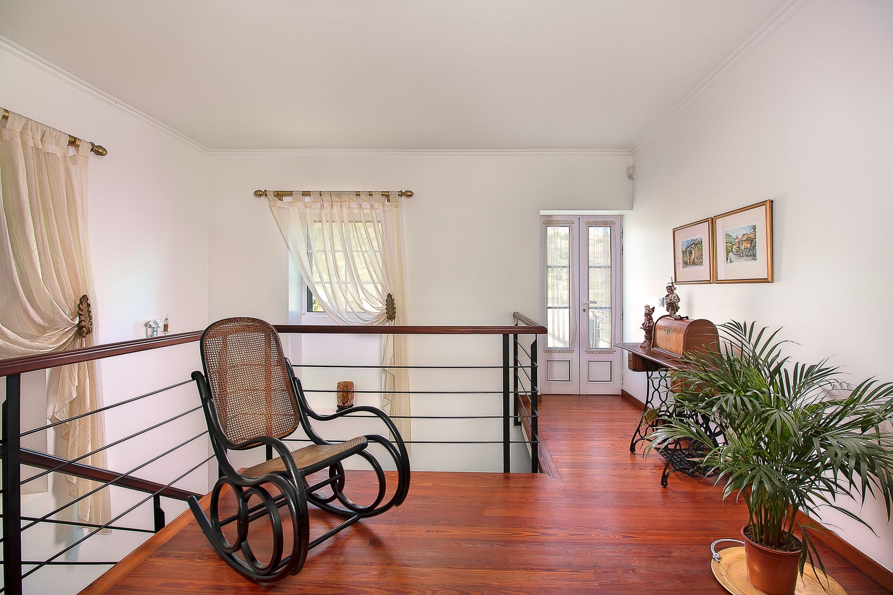 A spacious room with a wooden floor, a rocking chair, a potted plant, and a view of the outside through a door.