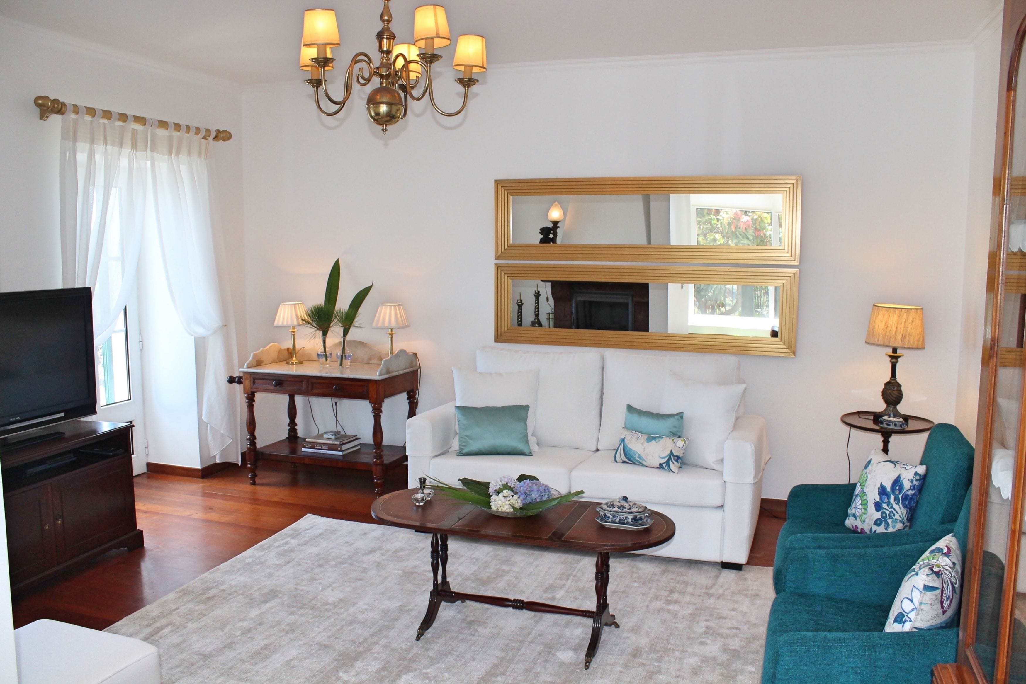 A living room with a television, a wooden coffee table, a white sofa, a teal armchair, and a chandelier.