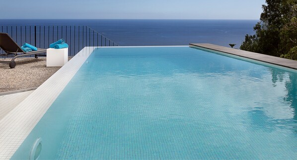 Ein klarer, blauer Infinity-Pool mit Blick auf den Ozean, eine Liege und ein weißes Podest.