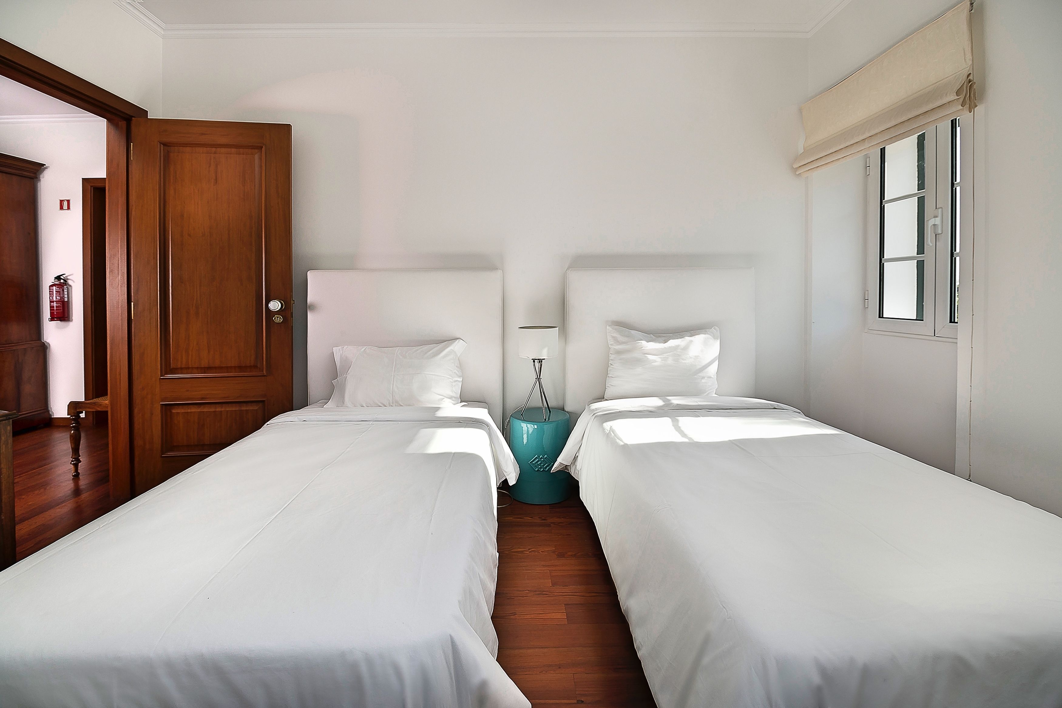 A hotel room with two single beds, a wooden door, a teal nightstand, and a window with a white blind.