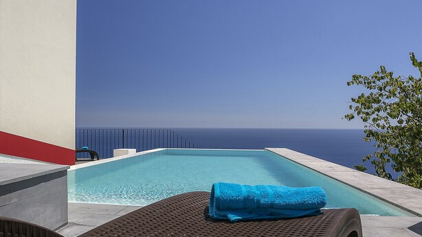 Una piscina con una toalla azul sobre una silla de descanso, con vista a un cielo azul despejado y al mar.