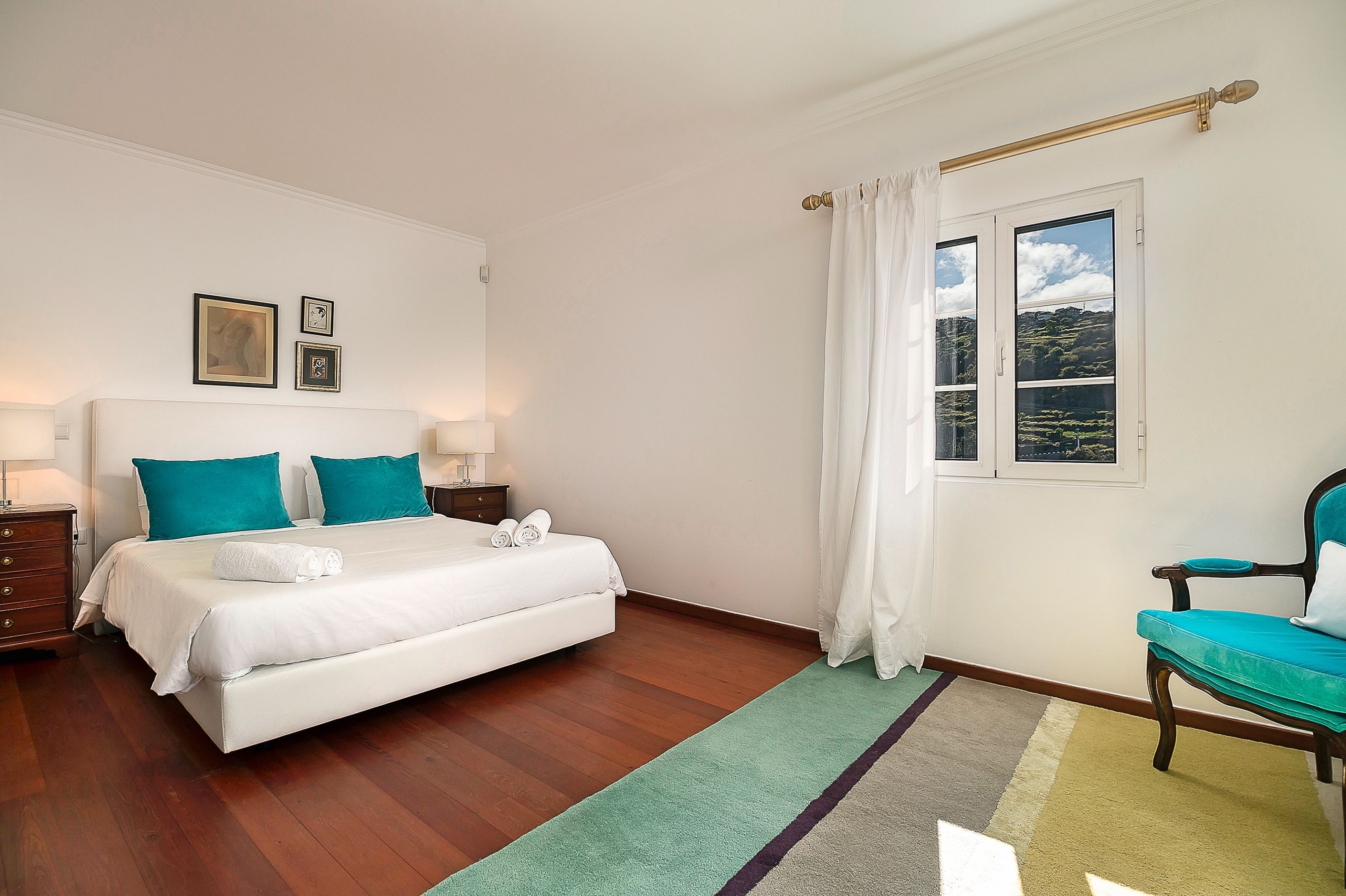 A bedroom with a bed, two bedside tables, a chair, a window with curtains, and a view of greenery outside.