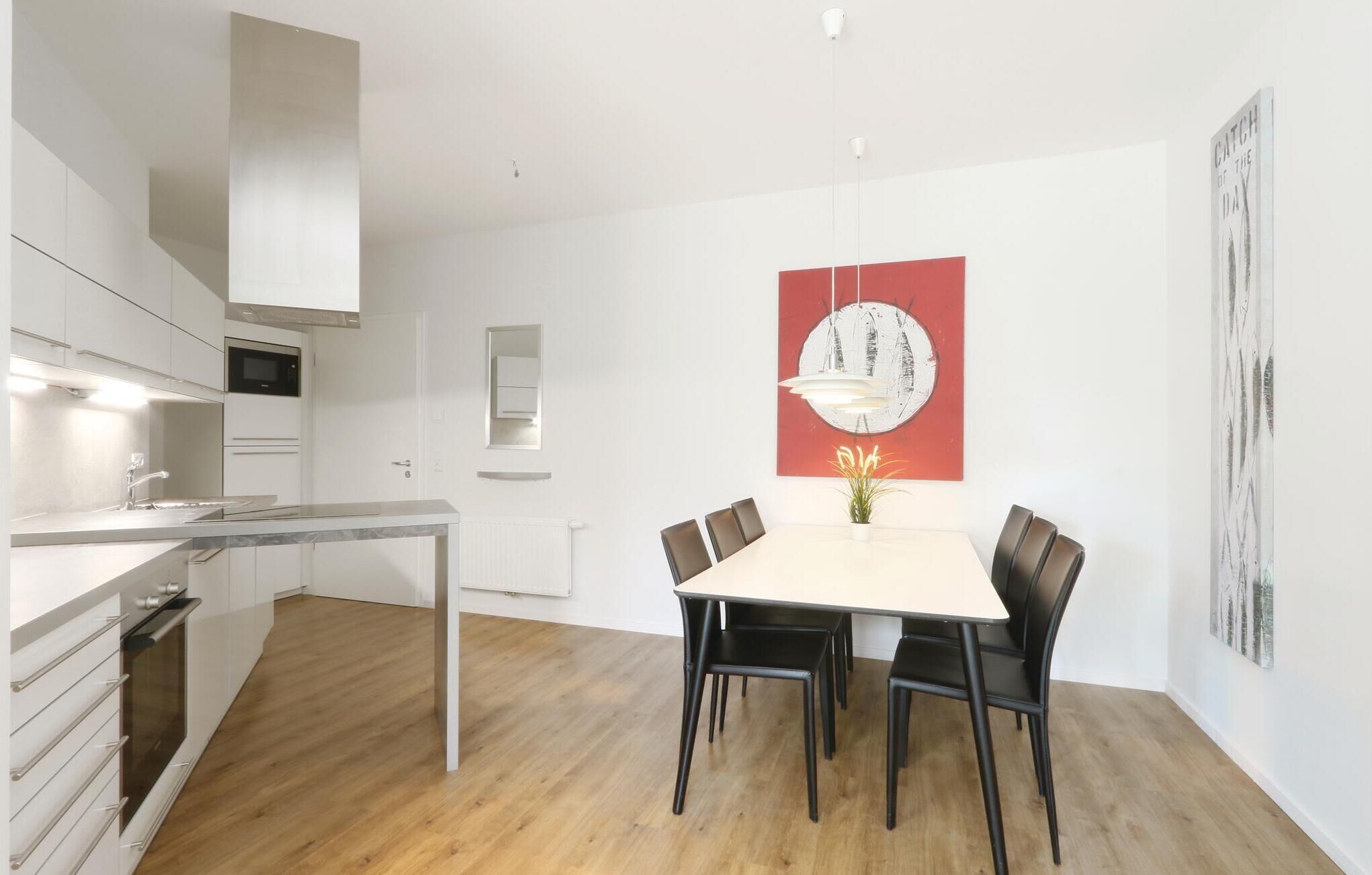 Cozinha moderna com armários brancos, forno embutido e micro-ondas. Há uma área de jantar com mesa branca e cadeiras pretas, e um quadro abstrato vermelho na parede.