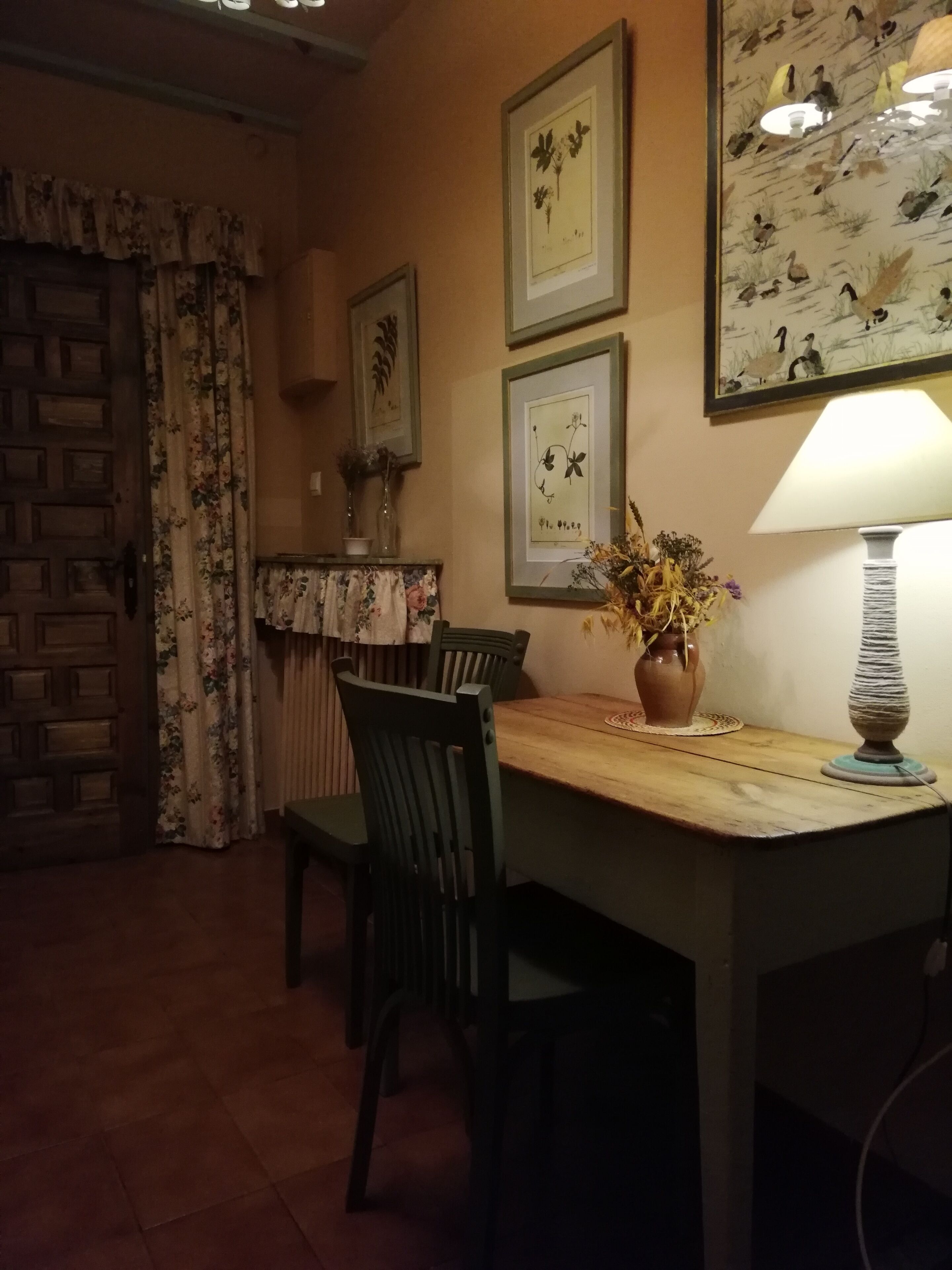 Un comedor con una mesa de madera, sillas y un florero con flores. En la pared hay grabados enmarcados con motivos botánicos.