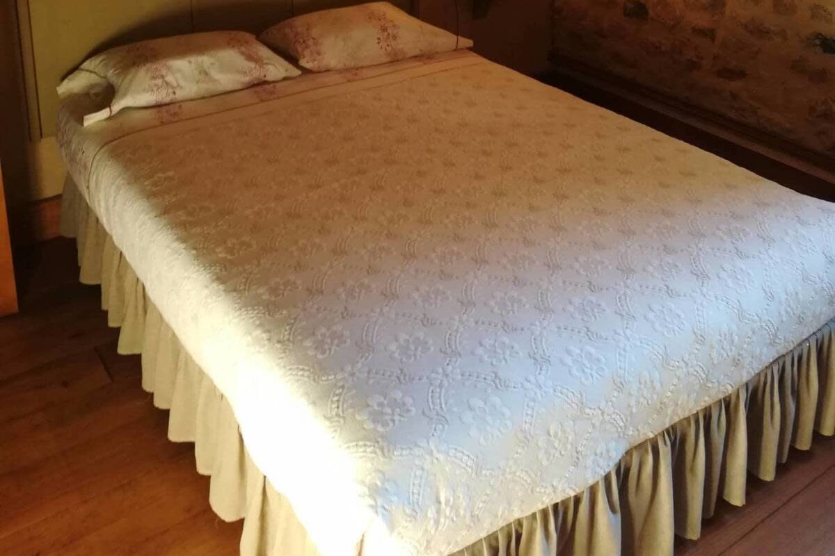 Un lit avec une couvre-lit à motifs et des oreillers, une table de chevet avec une lampe, un tableau encadré au mur et un mur en pierre.