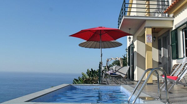 Uma área à beira da piscina com um guarda-sol vermelho, espreguiçadeiras e vista para o mar.